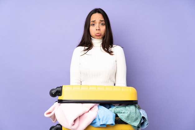 Photo voyageur jeune femme avec une valise pleine de vêtements sur un mur violet avec une expression triste et déprimée