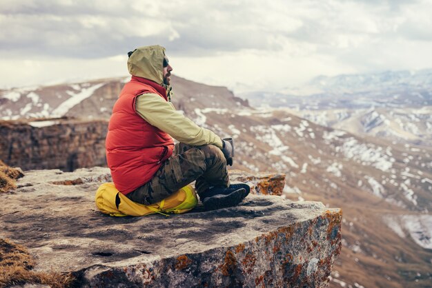Le voyageur masculin dans une veste rouge s'assied au bord de la falaise et apprécie la nature de montagne
