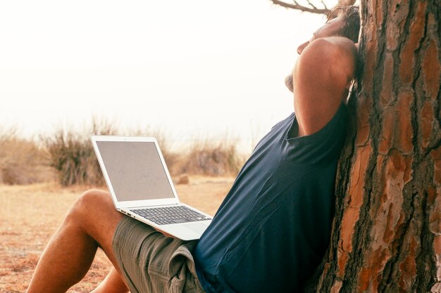 Un voyageur moderne après avoir travaillé sur un ordinateur portable, se détendant assis dans le parc contre un tronc d'arbre qui s'étire et profite de la liberté et du style de vie intelligent des nomades numériques.