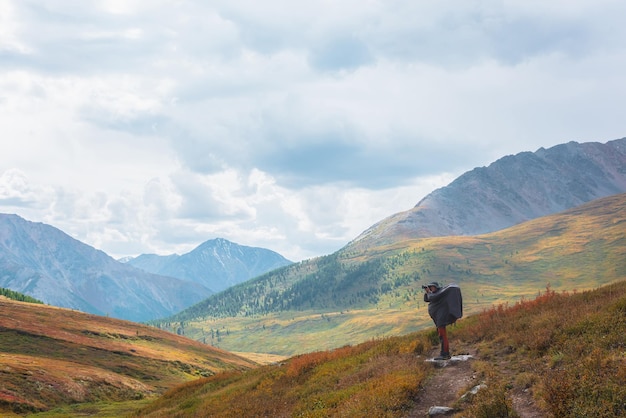 Voyageur seul en imperméable avec un grand sac à dos photographie la nature parmi les végétations d'automne qui s'estompent sur un sentier de randonnée à travers un col de montagne Rackpacker avec une caméra photo tourne un paysage de montagne d'autumn