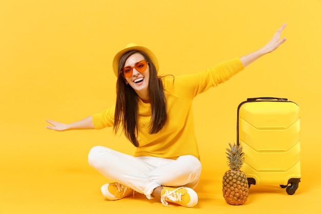Voyageur touriste femme en vêtements d'été chapeau écartant les mains, s'asseoir près de fruits ananas isolé sur fond jaune orange. Passager voyageant à l'étranger en week-end. Concept de voyage de vol aérien.