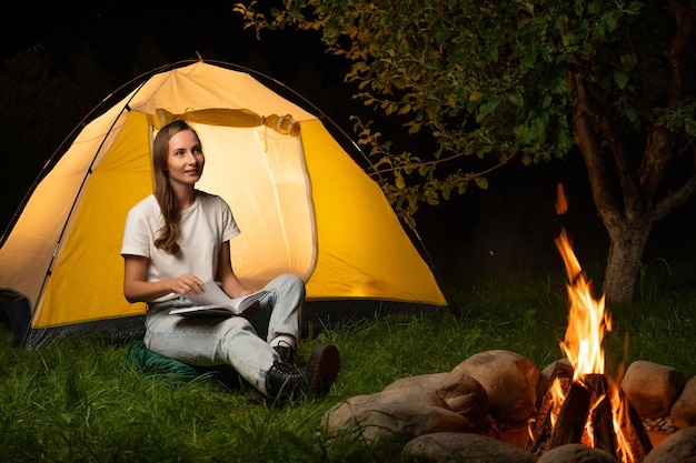 Une voyageuse campe dans un camping Une jeune femme est assise près de la tente et lit un livre dans le camp près du feu de camp