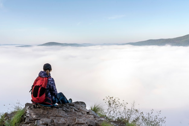Une voyageuse dans les montagnes est assise au bord d'une falaise et admire un matin brumeux dans le mo...