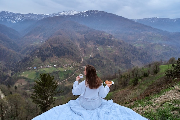 Une voyageuse portant un peignoir profitant d'un petit-déjeuner matinal et d'un paysage avec vue sur la montagne pendant la détente au lit. Moment de concept de voyage calme et tranquille où la personne ressent le bonheur et la liberté