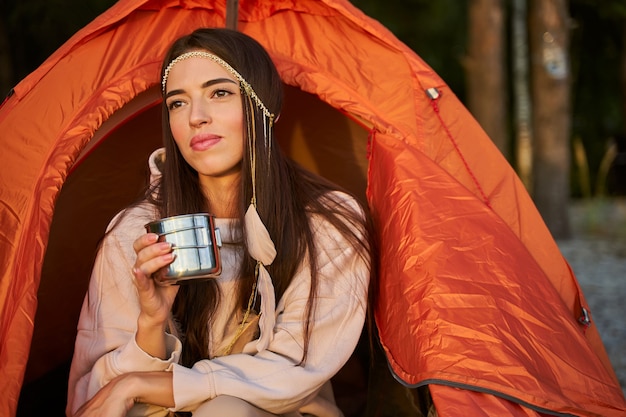 Voyageuse séduisante regardant loin et souriant tout en tenant une tasse de boisson chaude et assise à l'intérieur d'une tente de camp orange