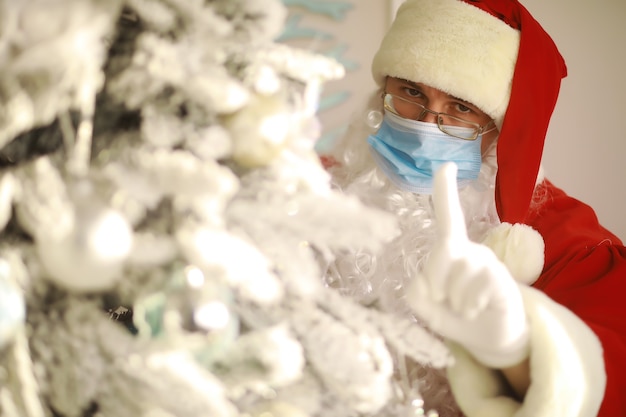 Vrai Père Noël et arbre de Noël sur fond, portant un masque de protection, des lunettes et un chapeau. Noël avec distance sociale. Covid-19