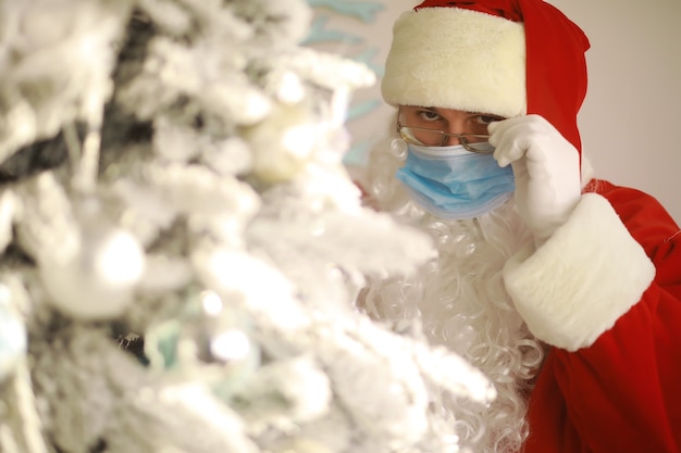 Vrai Père Noël et arbre de Noël sur fond, portant un masque de protection, des lunettes et un chapeau. Noël avec distance sociale. Covid-19