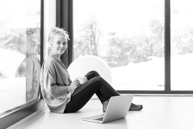 Vraie femme utilisant un ordinateur portable sur le sol, boire du café en profitant de la détente lors d'une froide journée d'hiver