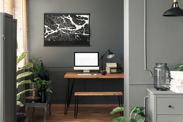 Vraie photo d'un intérieur de salon sombre et ouvert avec une affiche de carte sur le mur au-dessus d'un bureau en bois pour le travail à distance avec des maquettes de livres informatiques et une lampe