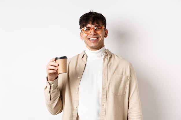 Vrais gens. Heureux jeune homme, boire du café dans une tasse en papier, commander des plats à emporter au café préféré, souriant, mur blanc.