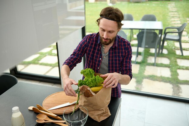 Vue aérienne d'un beau mec européen en chemise à carreaux debout sur la cuisine dans une villa spacieuse et déballant le pack écologique avec baguette de grains entiers, salades et légumes sains