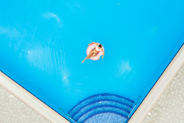 Vue aérienne de la belle jeune fille en bikini blanc reposant sur un beignet gonflable dans la piscine. Fond de vacances d'été