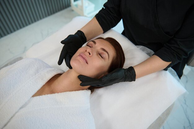 Vue aérienne d'une charmante élégante femme européenne d'âge moyen bénéficiant d'un massage professionnel sur son visage allongé sur un canapé pendant qu'une masseuse effectue un drainage lymphatique et un traitement facial anti-âge