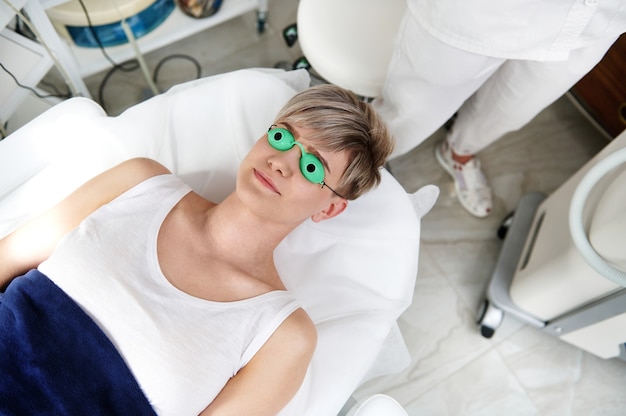Une vue aérienne d'un client d'une clinique de cosmétologie portant des lunettes de protection ultraviolettes allongé sur une table de massage avant une procédure d'épilation à l'aide d'une machine laser moderne