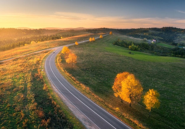 Vue aérienne des collines de la route des prairies vertes et des arbres colorés au coucher du soleil en automne Vue de dessus de la route rurale de montagne Beau paysage avec des arbres d'herbe verte de la chaussée avec un feuillage orange en automne