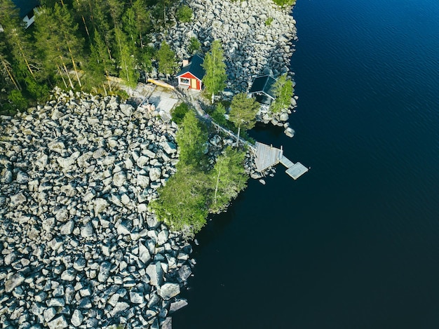 Vue aérienne de dessus d'une cabane en rondins rouge ou d'un chalet avec un sauna dans une forêt verte près d'un lac avec une côte rocheuse en Finlande