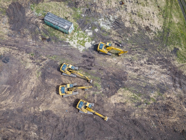 Vue aérienne de dessus de quatre pelles sur chenilles jaunes debout sur le sol près du chantier de construction et attendant la journée de travail pour commencer à creuser le sol près de la route de construction dans le champ parmi les arbres