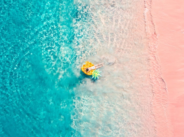 Vue aérienne de drone d'une fille flottant sur un ananas gonflable sur une plage rose tropicale exotique