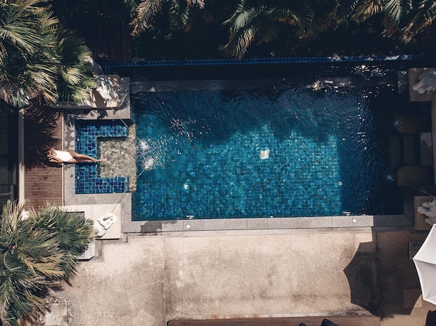 Vue aérienne du territoire de l'hôtel thaïlandais : toits de tuiles en terre cuite, palmiers, la piscine à l'eau bleu clair et une jeune femme s'y baignant ; drone.