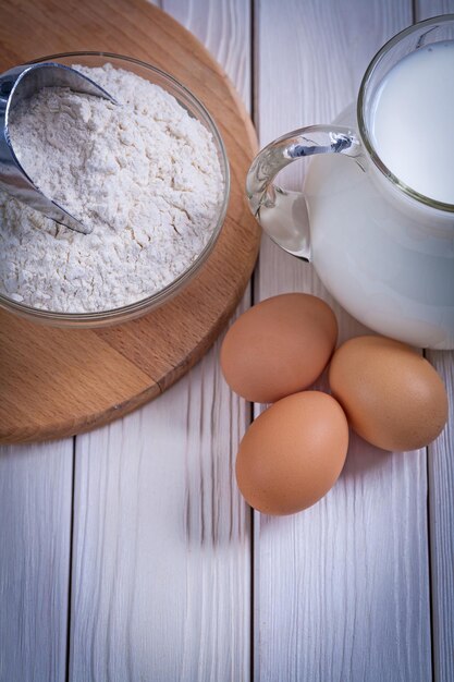 Vue aérienne farine dans un bol scoop oeufs pichet avec du lait sur une vieille planche de bois peinte en blanc concept de nourriture et de boisson