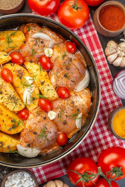 Vue aérienne de la farine de poulet cru avec pommes de terre légumes dans une casserole sur une serviette dépouillée rouge et tomates ails sur fond gris