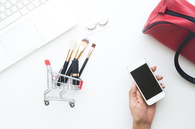 Vue aérienne de la femme tenant le téléphone mobile avec des objets de beauté dans le panier, concept en ligne Shopping