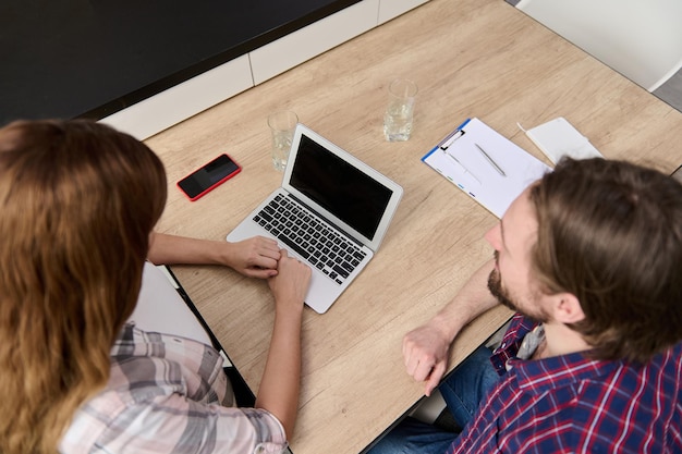 Vue aérienne d'une femme travaillant sur un ordinateur portable en tapant du texte sur le clavier assis à la table de la cuisine à côté de son mari souriant regardant l'écran du moniteur Achats en ligne et concept de travail à distance