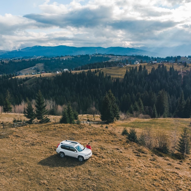 Vue aérienne de l'homme en manteau rouge près de la voiture suv blanche à la colline avec une vue magnifique sur les montagnes. voyage en voiture.