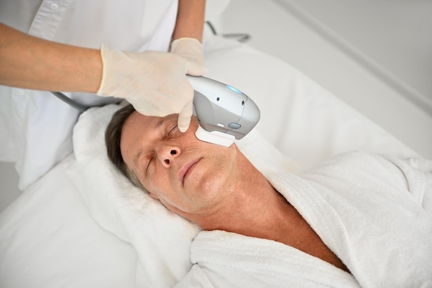 Photo vue aérienne d'un homme de race blanche mature recevant des soins de la peau masculins au centre de bien-être pendant que l'esthéticienne fait un traitement de beauté anti-âge sur son visage à l'aide d'un appareil médical à ultrasons