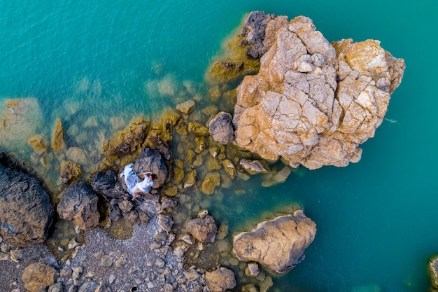 Vue aérienne d'une jeune femme vêtue d'une robe blanche sur les rochers. Paysage marin d'été avec une fille, plage, belles vagues, rochers, eau bleue. Vue d'en-haut. La nature