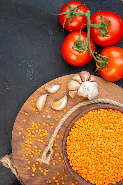 Vue aérienne de lentilles rouges dans un bol brun ails de corde sur planche ronde en bois et tomates avec tiges sur le côté gauche sur fond noir