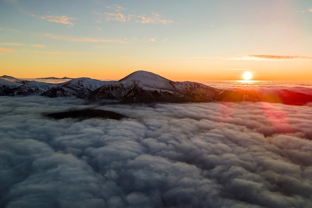 Vue aérienne d'un lever de soleil vibrant sur des nuages blancs et denses avec des montagnes sombres et lointaines à l'horizon.