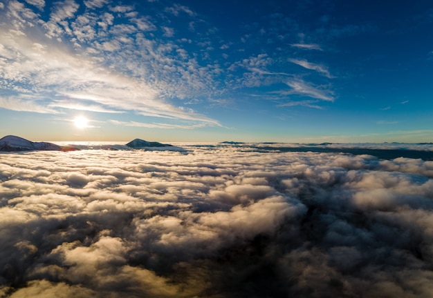 Vue aérienne d'un lever de soleil vibrant sur des nuages blancs et denses avec des montagnes sombres et lointaines à l'horizon.