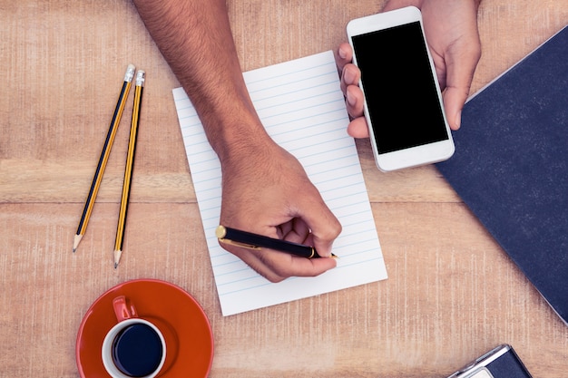 Vue aérienne de la main d&#39;homme d&#39;affaires sur le téléphone intelligent lors de l&#39;écriture sur le bloc-notes par la caméra et le café sur la table