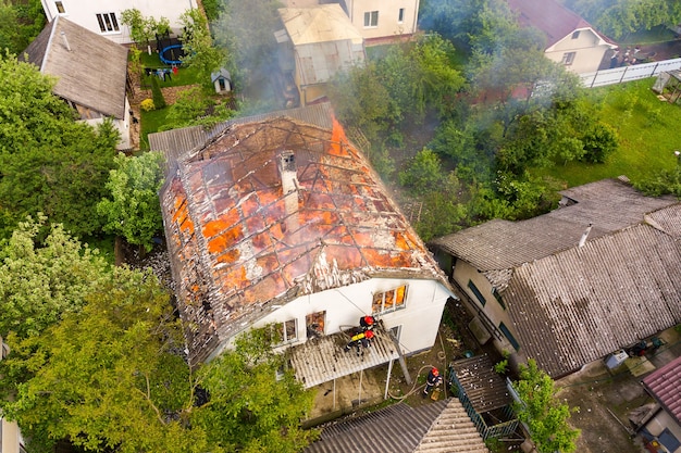 Vue aérienne d'une maison en feu avec des flammes oranges et une épaisse fumée blanche.