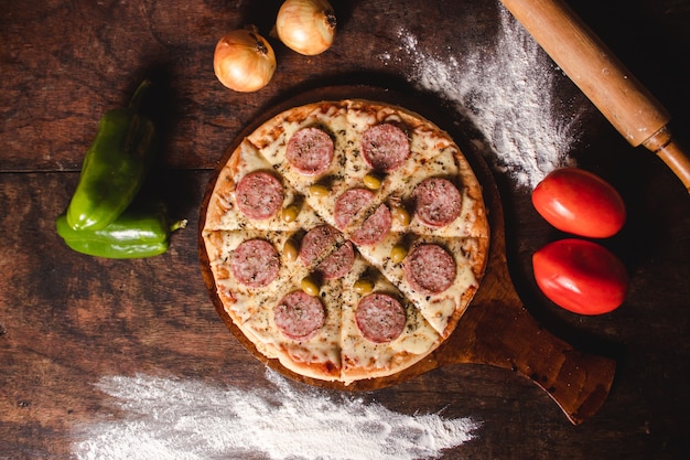 Vue aérienne d'une pizza au salami aux olives et au fromage avec des légumes autour sur une table en bois.