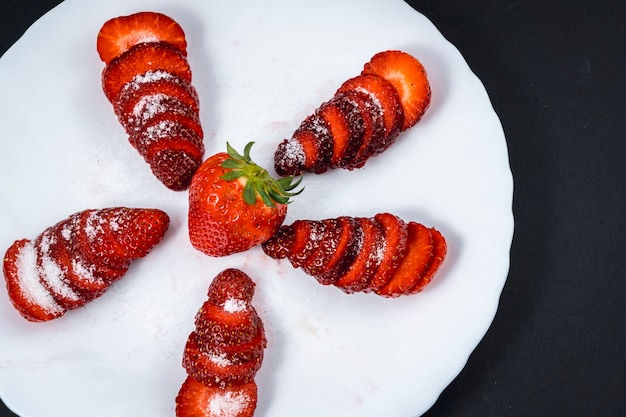 Vue aérienne de quelques fraises coupées avec du sucre sur une plaque blanche sur fond noir