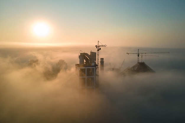 Vue aérienne de l'usine de ciment avec une haute structure de centrale en béton et une grue à tour sur un site de fabrication industrielle le soir brumeux. Concept de production et d'industrie mondiale.