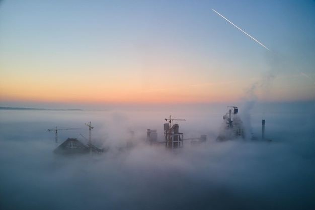 Vue aérienne de l'usine de ciment avec structure de centrale à béton élevée et grue à tour sur le site de fabrication industrielle le soir brumeux Production et concept industriel mondial