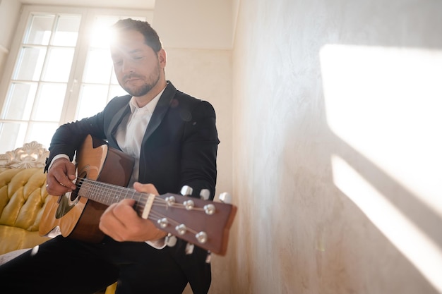 Photo vue à angle bas d'un beau jeune homme portant jouant de la guitare dans la chambre d'hôtel moderne avec la lumière du soleil le jour du mariage