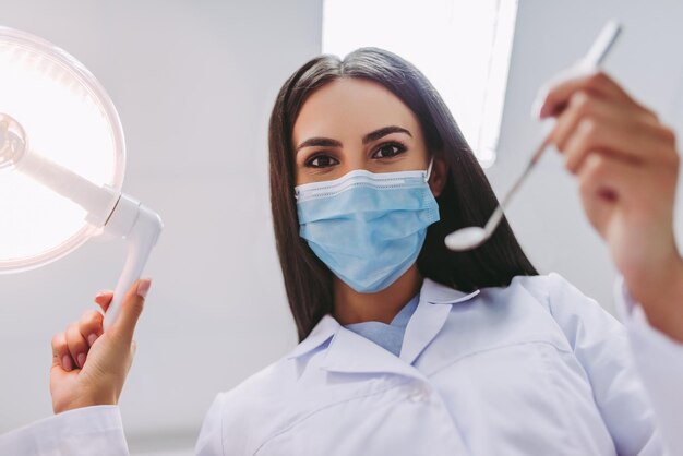 Vue en angle bas du dentiste en masque médical tenant des outils dentaires dans une clinique dentaire moderne