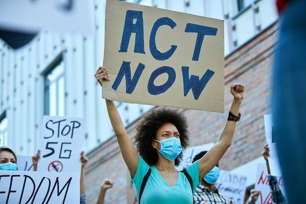 Vue en angle bas d'une femme afro-américaine portant un masque protecteur et portant une bannière avec l'inscription « agissez maintenant » lors de manifestations publiques