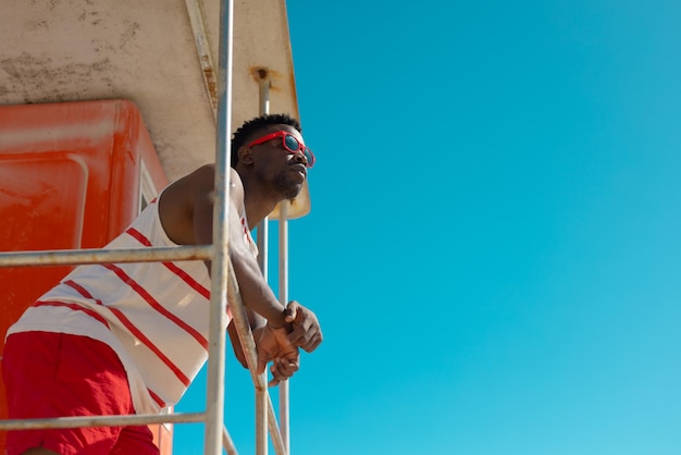 Vue à angle bas d'un jeune homme afro-américain à lunettes de soleil debout sur une cabane de maître nageur contre le ciel