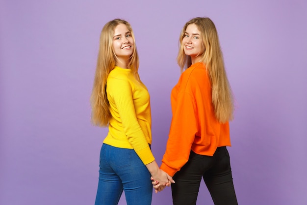 Vue arrière arrière de deux sœurs jumelles blondes souriantes dans des vêtements colorés se tenant la main, regardant en arrière isolées sur un mur bleu violet pastel. Concept de mode de vie familial de personnes.