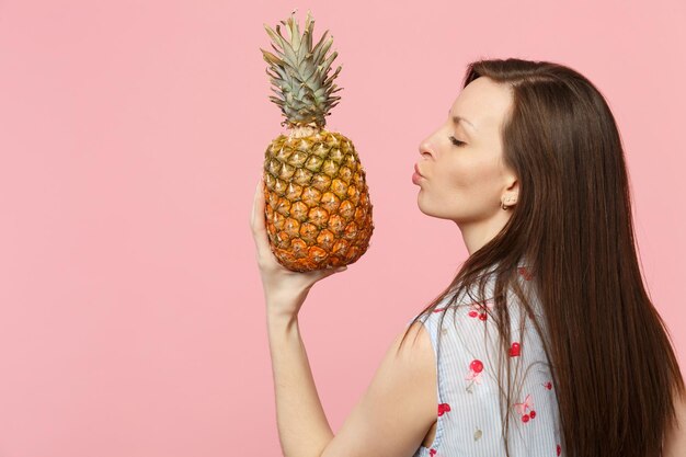 Vue arrière arrière d'une jeune fille regardant de côté soufflant en envoyant un baiser d'air à des fruits d'ananas mûrs frais isolés sur fond pastel rose. Les gens vivent un style de vie vivant et se détendent. Maquette de l'espace de copie.