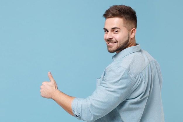 Vue arrière arrière d'un jeune homme barbu souriant de 20 ans en chemise décontractée posant isolé sur fond bleu pastel en studio. Concept de style de vie des gens. Maquette de l'espace de copie. Montrant les pouces vers le haut, regardant la caméra.