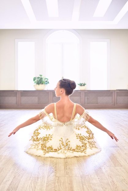 Vue arrière de la belle ballerine en robe crème assise et se réchauffe les mains sur le parquet dans le studio de ballet