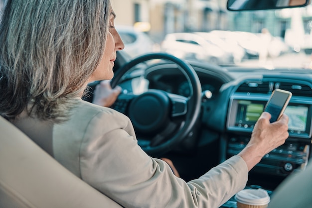 Vue arrière d'une belle femme mature en tenue décontractée intelligente à l'aide d'un téléphone en conduisant une voiture