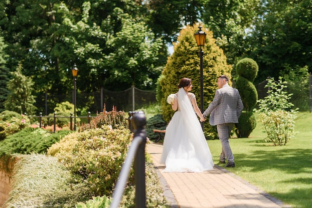 Vue arrière de la belle mariée en robe blanche et du marié élégant en costume qui se tient la main belle se regardant tout en