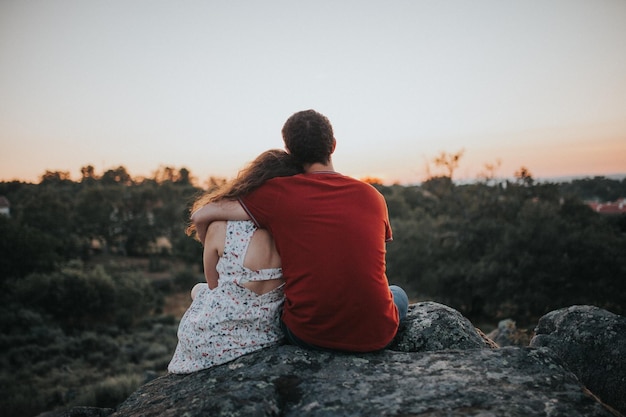 Vue arrière d'un couple assis sur un rocher contre un ciel clair au coucher du soleil